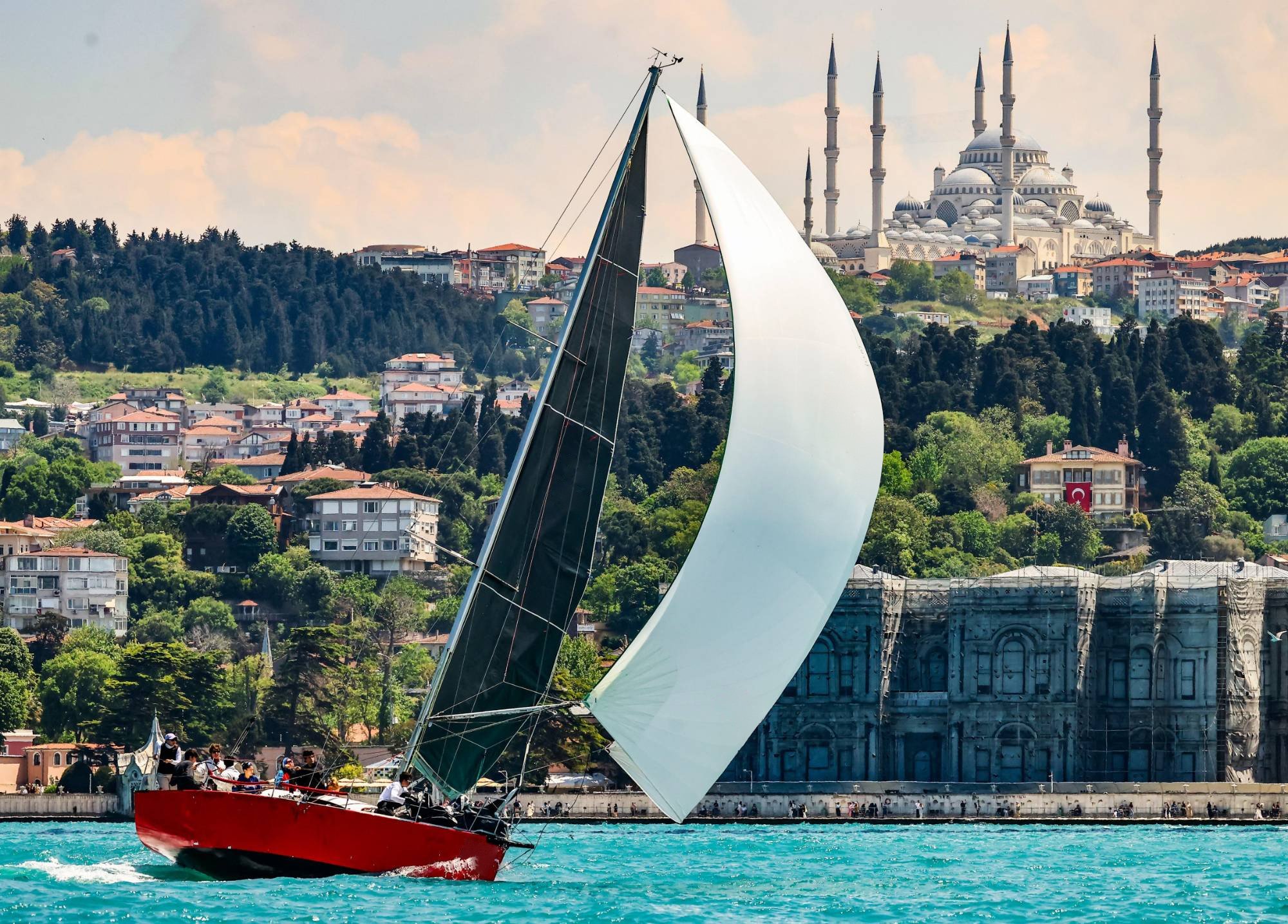 Kırmızı gövdeli bir yarış teknesi, arkasında Çamlıca Camii ve Boğaz kıyısıyla beyaz balon yelkeniyle seyrediyor.
